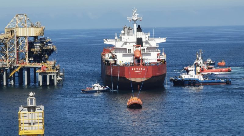 Granelero amarrado a una boya frente a una plataforma de carga, asistido por dos remolcadores y una lancha de servicio.