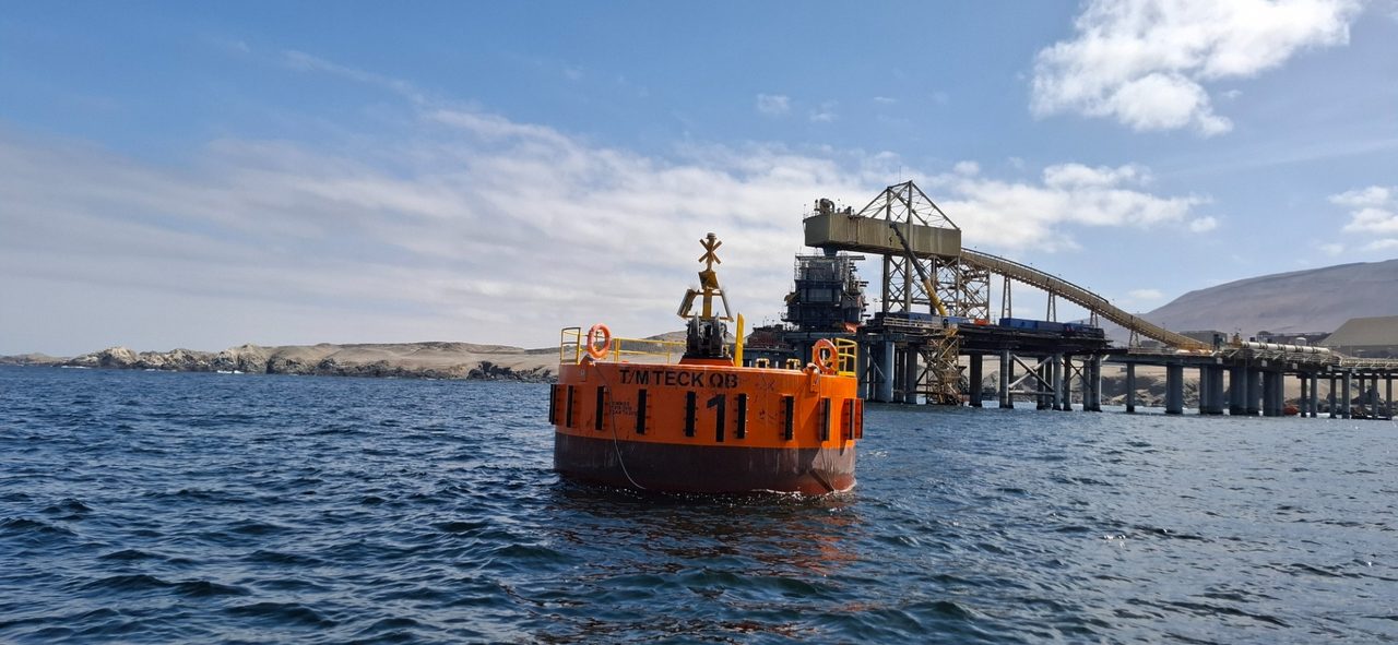 Boya de amarre cilíndrica de acero fondeada frente al muelle de carga de un terminal marítimo industrial del norte de Chile, con la correa transportadora y el cerro costero al fondo.